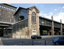 Historischer S-Bahnhof mit einer Fassade aus Glas und Stein im spätklassizistischen Stil, einer großen Treppe im Vordergrund und erhöhten Gleisen, die von einer Metallkonstruktion getragen werden. Die Straße und parkende Autos befinden sich im Vordergrund, während der Bahnhof von modernen Gebäuden umgeben ist.