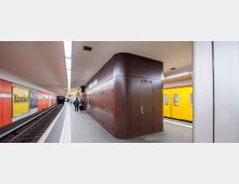 U-Bahn-Station mit zwei gegenüberliegenden Bahnsteigen, einer gelben U-Bahn im Hintergrund und orangefarbenen Wandverkleidungen. Ein zentraler brauner Technikraum trennt die beiden Gleise, an denen wenige Personen stehen.