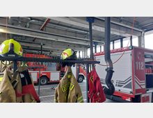 Innenansicht einer Feuerwache mit roten Einsatzfahrzeugen im Hintergrund, aufgehängter Feuerwehrschutzkleidung und Helmen auf einer Kleiderstange im Vordergrund. An der Decke sind Abluftschläuche für die Fahrzeugabgase angebracht.