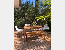 Holzterrasse mit einem Gartenmöbel-Set aus Holz, bestehend aus einem Tisch, einer Bank und weißen Stühlen, umgeben von dichtem grünen Bewuchs und Bäumen. Eine Vase mit rosa Tulpen steht auf dem Tisch, im Hintergrund ist blauer Himmel sichtbar.