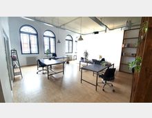 Helles Büro mit hohen Decken, großen Rundbogenfenstern und hellem Holzfußboden. Der Raum ist mit mehreren Tischen, Bürostühlen, Pflanzen und Regalen ausgestattet.