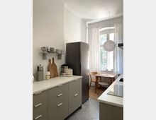 Minimalistische Küche mit grauen Unterschränken und weißer Arbeitsplatte, daneben ein hoher, dunkler Kühlschrank. Im Hintergrund ein Essbereich mit Holztisch und Stühlen vor einem großen Fenster mit weißen Vorhängen; eine runde Papierlampe hängt von der Decke.