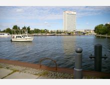 Uferbereich mit Kopfsteinpflaster und Pollern im Vordergrund, ein Fluss mit einem Boot und weiteren Schiffen im Wasser, im Hintergrund ein Hochhaus mit der Aufschrift "Hotel Mercure" sowie Bäume und Gebäude entlang der Promenade.
