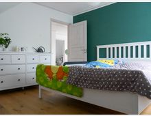 Schlafzimmer mit einem weißen Bett und einer gemusterten Bettdecke, daneben eine weiße Kommode mit Pflanzen und Dekoration. Die Wände sind in Weiß und Dunkelgrün gehalten, der Holzboden ist hell, und eine offene Tür führt in einen angrenzenden Raum.