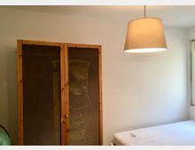 Ein minimalistisches Schlafzimmer mit einem Holzschrank auf der linken Seite, einer Matratze ohne Bettgestell rechts und einer Hängelampe mit beigem Schirm. Die Wände sind weiß, und ein Fenster lässt Tageslicht herein.