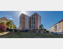 Zwei hohe, quadratische Wohnblocks mit braunen und weißen Fassaden, eingefasst von einer Grünfläche im Vordergrund. Rechts sind mehrstöckige Altbauten mit geparkten Autos und links einzelne Bäume zu sehen, bei klarem Himmel im Hintergrund.