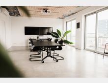 Moderner Besprechungsraum mit einem langen schwarzen Tisch, umgeben von schwarzen Stühlen. Der Raum hat große Fenster mit Blick auf eine Stadtlandschaft, eine grüne Pflanze in der Ecke und eine Decke mit einem dekorativen Holzmuster und einem Projektor.