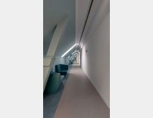 Langer, schlanker Korridor mit schrägen Decken, weißen Wänden und einer durchgehenden Lichtleiste an der Decke. Auf der linken Seite sind moderne Sitzgelegenheiten in dunklen Farben entlang der Wand angeordnet.