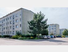 Mehrfamilienhäuser aus hellem Putz mit mehreren Stockwerken, umgeben von Grünflächen mit Bäumen und Blumenbeeten. Im Vordergrund befindet sich ein Parkplatz mit abgestellten Autos.