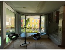 Ein moderner Innenraum mit Betonwänden, einer kleinen Wendeltreppe und einem großen Fenster, das auf einen Balkon mit Pflanzen und dahinterliegende bunte Wohnhäuser blickt. Zwei schwarze Stühle und ein runder Tisch befinden sich vor dem Fenster auf einem Holzboden.