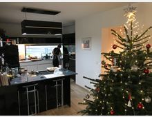 Moderne Wohnküche mit dunkler Kücheninsel und Barhockern, schwarzen Hängelampen sowie einer langen Fensterfront über der Arbeitsfläche. Rechts steht ein großer, mit Lichterkette, Kugeln und Sternspitze geschmückter Weihnachtsbaum vor einer weißen Wandnische.