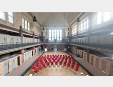 Eine historische Schwimmhalle mit hohen Decken, großen Fenstern und einer umlaufenden Galerie. Im Beckenbereich stehen rote Stühle in einem Kreis auf Holzfußboden, während die Umkleidekabinen an den Wänden erhalten geblieben sind.