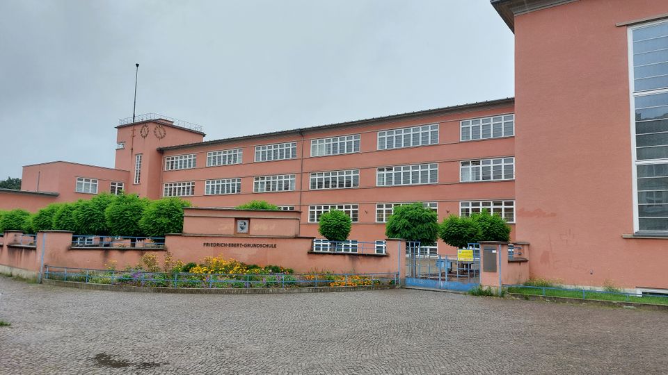 Rosa Schulgebäude der Friedrich-Ebert-Grundschule mit mehreren rechteckigen Fenstern, einer zentralen Turmstruktur mit Uhr und umliegenden gepflegten grünen Bäumen und Blumenbeeten. Im Vordergrund ein gepflasterter Platz und ein blaues Metalltor.