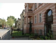 Straßenszene mit einer Reihe historischer, mehrstöckiger Gebäude, darunter ein rosa Gebäude mit Balkonen und hohen Fenstern. Im Vordergrund ein schmiedeeiserner Zaun, dahinter kleine Grünflächen und Laternen im klassischen Stil; links ist eine gepflasterte Straße mit parkenden Autos und Bäumen zu sehen.