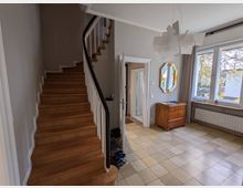 Heller Eingangsbereich eines Wohnhauses mit geschwungener Holztreppe und schwarzem Handlauf, beigen Fliesen sowie weiß-grauen Wänden. Rechts befinden sich große Fenster mit Vorhängen, ein Heizkörper, eine Holzkommode und ein achteckiger Wandspiegel; in der Mitte führt eine Tür in den Flur.