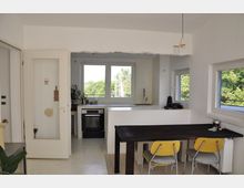 Wohnküche mit weißen Wänden und großen Fenstern, die Blick auf grüne Bäume bieten. Ein dunkler Holztisch mit gelben Stühlen steht zentral im Raum, während im Hintergrund eine Küche mit Herd, Spüle und Arbeitsflächen zu sehen ist.