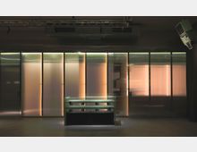 Dunkler Innenraum mit einer Wand aus transluzenten, beleuchteten Paneelen, die warmes Licht abstrahlen. Im Vordergrund steht ein kleiner Pulttisch mit einer Gitterstruktur und sichtbaren Lautsprechern.