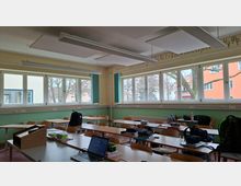 Klassenzimmer mit mehreren Reihen aus Holztischen und -stühlen sowie einer Lehrerpult-Ecke im Vordergrund. Entlang der Wand befinden sich breite Fensterfronten mit Blick auf Bäume und Wohngebäude; an der Decke hängen lange Leuchtstofflampen und Akustikplatten.