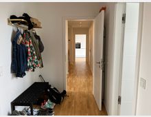 Flur in einer Wohnung mit hellem Holzfußboden, an der linken Wand befindet sich eine Garderobe mit mehreren Jacken und ein schwarzes Schuhregal mit Schuhen darunter. Im Hintergrund führt der Flur zu einer offenen Küche mit weißen Schränken.