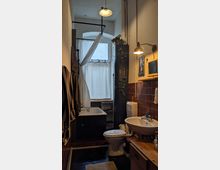 Schmales Badezimmer mit Badewanne, WC und Waschbecken auf einer Seite. Die Wände sind teilweise mit dunkelbraunen Fliesen verkleidet, und ein kleines Fenster mit weißem Vorhang lässt Licht herein. Deckenlampen und eine hängende Pflanze sorgen für Dekoration.