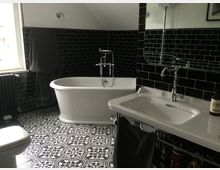 Ein Badezimmer mit einer freistehenden weißen Badewanne, einem Waschbecken mit Spiegel und einem dunklen Fliesenboden im Ornamentstil. Die Wände sind mit glänzenden schwarzen Kacheln gefliest, und ein Fenster mit hellem Tageslicht befindet sich links.
