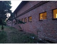 Langes Gebäude aus rotem Backstein mit einer Reihe kleiner, rechteckiger Fenster, durch die warmes Licht nach außen scheint; im Vordergrund Gras und vereinzelte Büsche, daneben ein Nadelbaum.