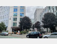 Straßenszene mit modernen Wohn- und Geschäftshäusern, großen Fenstern und Bäumen entlang des Gehwegs. Im Vordergrund sind parkende Autos, ein Café mit Außensitzplätzen und eine Fußgängerin sichtbar.