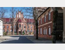 Straßenszene mit einem alten, roten Backsteingebäude im neogotischen Stil, das spitze Bögen und Ziergiebel aufweist. Im Vordergrund stehen Bäume und ein Gehweg, während der Himmel klar und blau ist.