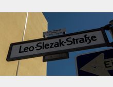 Straßenschild mit der Aufschrift „Leo-Slezak-Straße“ vor blauem Himmel und einem hellgelben Gebäude im Hintergrund. Darüber ein kleineres Schild mit der Beschriftung „Leo Slezak, Sänger, *1873 †1946“.