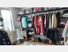 Offenes Ankleidezimmer mit freistehendem Metall-Kleiderregal, an dem zahlreiche Kleidungsstücke auf Bügeln hängen, sowie Ablagefächern mit gefalteter Wäsche, Schuhreihen und Aufbewahrungsboxen. Daneben stehen ein großer Standspiegel und ein Fenster mit Jalousie in einem hellen Raum mit Holzboden.