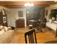 Ein Wohnzimmer mit Holzboden und sichtbaren Deckenbalken. Im Raum befinden sich ein Sofa mit Hund, ein Regal mit Büchern, ein Schreibtisch mit Computer sowie ein Schlagzeug und einige Pflanzen auf der Fensterbank.
