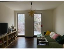 Wohnzimmer mit dunklem Holzboden, grünem Ecksofa und Couchtisch, links ein Fernseher auf einem niedrigen Regal. Zwei bodentiefe Fenster/Terrassentüren geben den Blick in einen Garten frei, an der Decke hängt eine sternförmige Pendelleuchte.