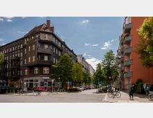 Straßenecke in einer urbanen Umgebung mit mehrstöckigen Wohngebäuden; links ein dunkelgraues Eckhaus mit Ladenlokalen im Erdgeschoss, rechts ein orangefarbenes Gebäude mit Balkonen. Bäume säumen die Straße, und am Gehweg sind Fahrräder abgestellt.