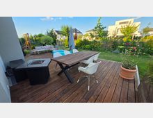 Holzterrasse in einem Wohnhausgarten mit großem Esstisch und weißen Stühlen, Feuerstelle, Loungesofa sowie Sonnenschirm und kleinem Kinderpool. Dahinter liegen Rasen, Blumenbeete und Bäume; im Hintergrund sind moderne Einfamilienhäuser zu sehen.