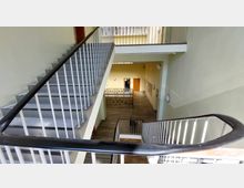 Treppenhaus eines mehrstöckigen Gebäudes mit Metallgeländern und hellem Anstrich. Im Hintergrund sind eine Holztür, ein Geländer mit dekorativen Elementen und ein Holzfußboden zu sehen.