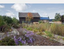 Ein Garten mit blühenden violetten und gelben Blumen, umgeben von Sträuchern und Pflanzen, im Vordergrund ein dekoratives hölzernes Wagenrad. Im Hintergrund ein dunkles Holzgebäude mit ziegelgedecktem Dach, seitlich moderne Gebäude mit blauen Dächern.