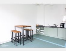 Helle Küche mit mintgrünen Unterschränken, einer schwarzen Arbeitsplatte, Spüle, Wasserkocher, Kaffeemaschine und offenem Regal mit Geschirr. Links steht ein hoher Holztisch mit vier passenden Hockern auf einem türkisfarbenen Boden.