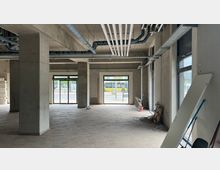 Innenraum eines unfertigen Gebäudes mit freiliegenden Betonwänden, sichtbaren Rohren und Kabeln an der Decke sowie großen bodentiefen Fenstern, die Tageslicht hereinlassen. Im Vordergrund befinden sich Baumaterialien und Werkzeuge, der Boden ist bereits mit Fliesen versehen.