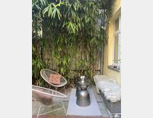 Kleiner, gemütlicher Innenhof mit Bambuspflanzen als Hintergrund, zwei runden Stühlen mit Kissen, einem kleinen schwarzen Beistelltisch mit Blumendekoration und einer Sitzbank mit Fellauflage an einer gelben Hauswand.