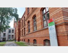 Rotes Backsteingebäude mit großen Rundbogenfenstern, verziert mit helleren Ziegelmustern; im Vordergrund ein Weg, daneben ein Schild mit einem Lageplan und der Aufschrift „Zentrale Aufnahme und Diagnostik (ZAD)“. Links ist ein moderner Anbau mit weißer Fassade zu sehen.