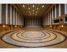 Ein großer Konzert- oder Veranstaltungssaal mit Holzvertäfelung, einer abgerundeten Bühne mit konzentrischen Linien und erhöhten Sitzreihen im Hintergrund. Die Wände sind mit hohen Fensterbereichen und Gittermustern gestaltet, die Decke ist mit quadratischen Paneelen verkleidet, und seitlich hängt eine große Uhr.