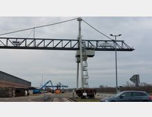 Hafenbereich mit einem großen, metallenen Portalkran, beschildert mit „Stadthafen Brandenburg a.d.H.“. Im Hintergrund sind Stapel von Baumaterialien, ein Lagergebäude sowie Arbeitsmaschinen sichtbar.