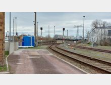 Bahngelände mit Gleisen und Signalanlagen. Im Hintergrund stehen Güterwaggons auf einer Brücke, daneben befinden sich ein blaues Gebäude und ein weißes Industriegebäude am rechten Bildrand, umgeben von kahlen Bäumen und Freiflächen.