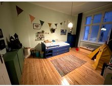 Kinderzimmer mit hellem Holzboden, einem niedrigen blauen Bett mit Schubladen, einem bunten Teppich und einer gelben Zeltspielhöhle. An der Wand hängen dreieckige Wimpelgirlanden, Regale und Dekorationen, und ein großes Fenster lässt blaues Abendlicht herein.