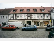 Fachwerkhaus mit rot gedecktem Dach und mehreren Gaubenfenstern, im Erdgeschoss verschiedene Ladengeschäfte. Davor eine gepflasterte Straße mit parkenden Autos und einem Lieferwagen.