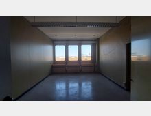 Ein leerer Raum mit beigen Wänden, einer Decke mit weißen Akustikplatten und einem glänzenden Boden. Am Ende des Raumes befinden sich drei große Fenster, die Tageslicht hereinlassen und einen Blick auf eine Außenszene mit Dächern und Bauwerken bieten.