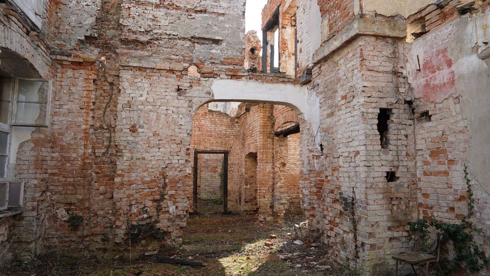 Innenhof einer verfallenen Backsteinruine mit bröckelndem Putz, offenen Fenster- und Türöffnungen sowie einem gemauerten Rundbogen-Durchgang. Der Boden ist mit Moos und Schutt bedeckt, und durch das fehlende Dach fällt Tageslicht in den Raum.