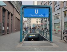 Straßeneingang zur U-Bahn-Station „Friedrichstraße“ in Berlin, gekennzeichnet durch ein großes blaues Schild mit weißem „U“. Umgeben von Gebäuden mit roten Ziegel- und Glasfassaden, Fußgängerverkehr und Geschäften im Hintergrund.