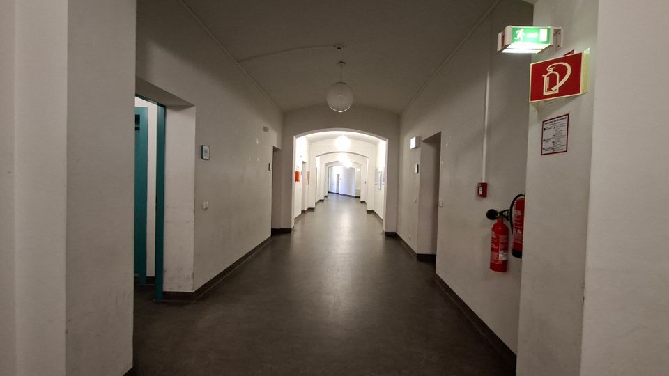 Langer, weiß gestrichener Innenflur mit gewölbter Decke und runden Deckenleuchten, entlang der Wände mehrere Türöffnungen und Durchgänge mit Bögen. An der rechten Wand befinden sich ein Feuerlöscher sowie ein grünes Notausgangsschild und ein rotes Brandschutzsymbol.