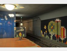 U-Bahn-Station mit rot gefliesten Böden, blauen Wandpaneelen und dekorativen Mosaiken mit pflanzlichen Mustern an der Wand. Im Hintergrund sind die Gleise und ein Signal mit grünem Licht zu sehen.